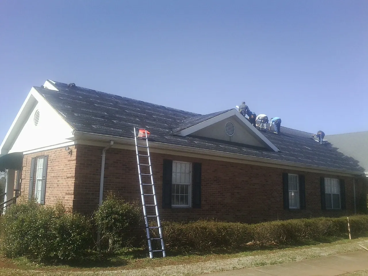 Crew tearing shingles off a brick ranch roof with a ladder set — Emergency Roof Repair work by our crew in Genesee